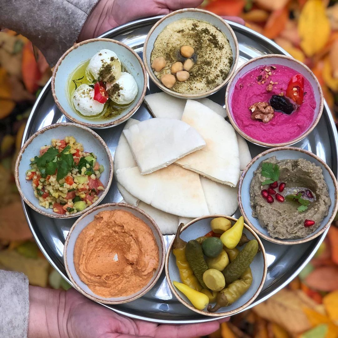 A platter with slices of flatbread in the middle surrounded by small bowls of various dishes, including boiled eggs, bright beet dip, hummus with chickpeas, pickles, baba ganoush, and chopped salad