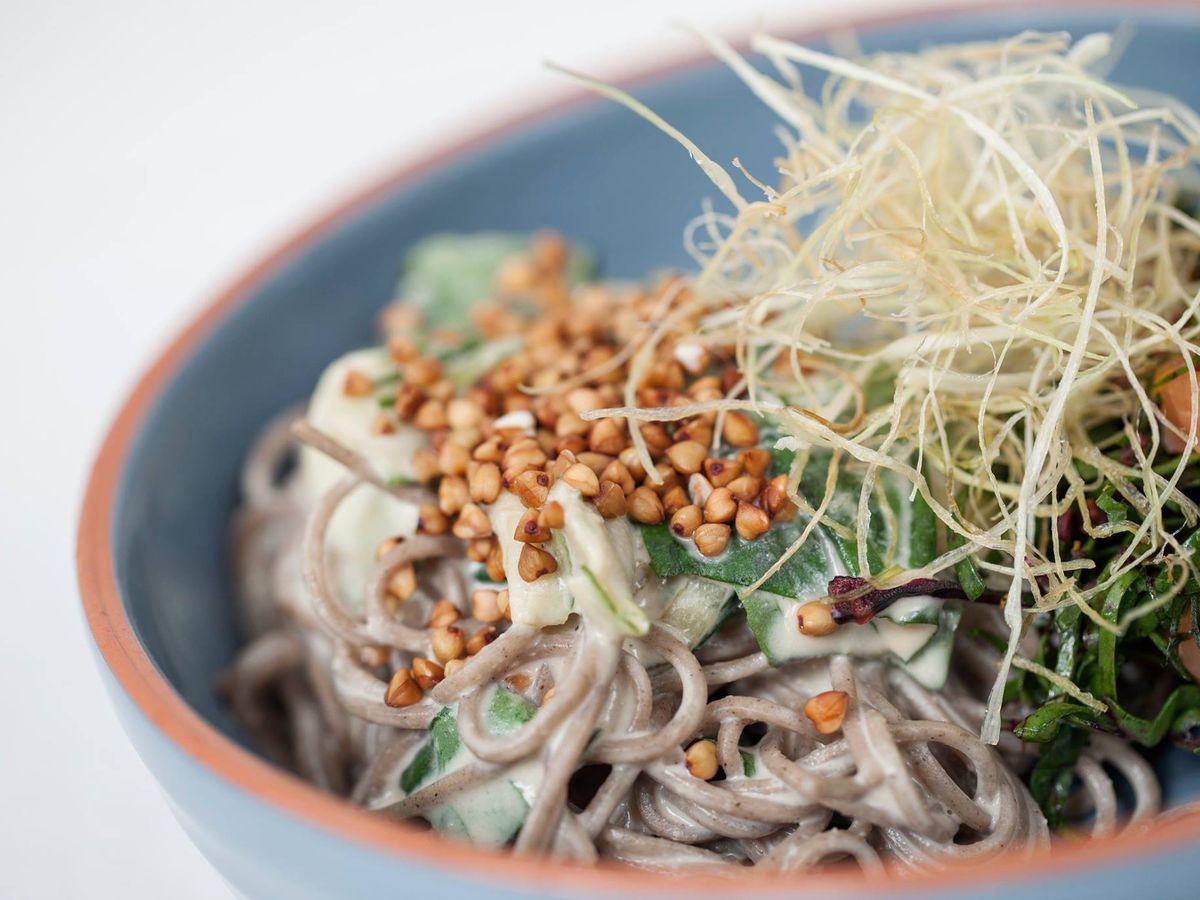 A bowl of soba noodles on a blank white background. The noodles are tossed in a white tahini sauce, and topped with pieces of buckwheat and a nest of shaved vegetables