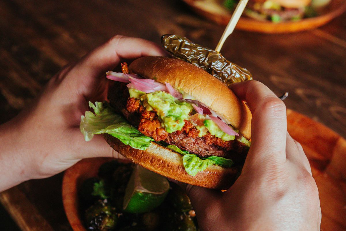 Two hands holding a burger with a chile speared on top over a red basket