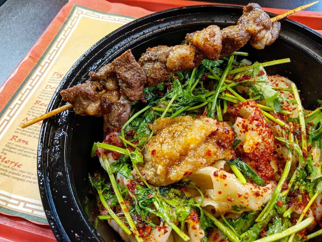 A black plastic bowl of thick hand-pulled noodles, heavily dusted with chile powder and topped with greens and a generous dollop of garlic. A wooden skewer of lamb pieces sits across the rim of the bowl, which is on a Chinese Zodiac placemat on a red tray