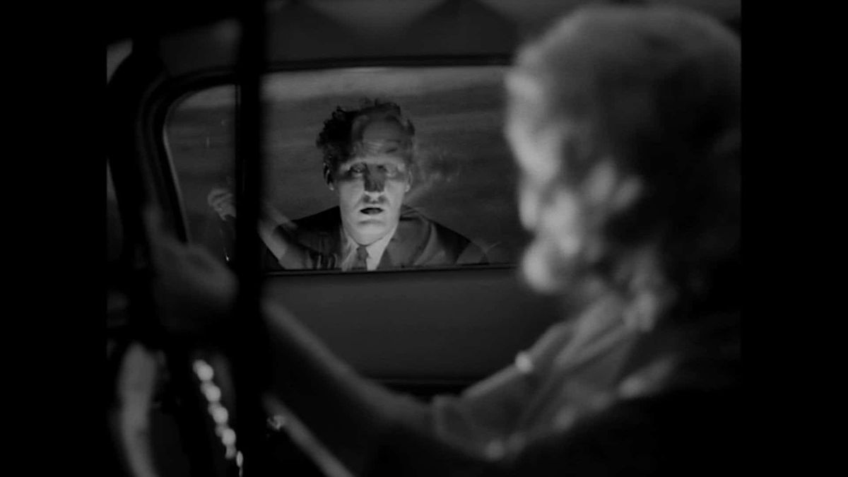 A man peers into a car window as a woman looks on from the driver’s seat. The image is in black and white.