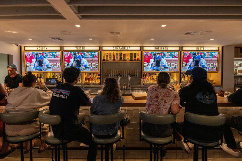 A diverse group of young diners sits on plush bar stools, facing a row of TV screens displaying a baseball game.