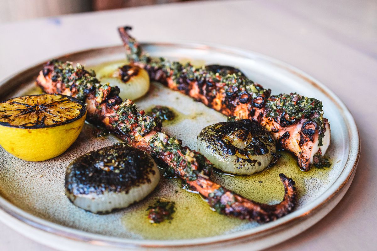 A stoneware plate with two tentacles of grilled octopus and charred lemon and onions and a puddle of olive oil