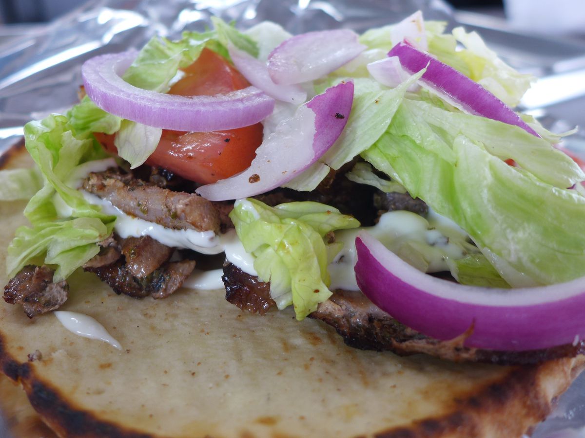 A flat pita with grilled pork, lettuce, and purple onions piles high with white sauce.