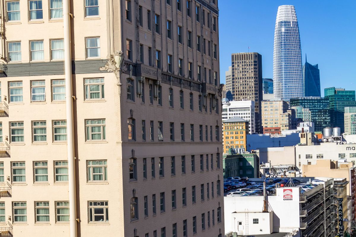 An extremely high-angle shot of a the side of a tall, sandstone-colored high-rise building in San Francisco, with the blue, obelisk-shaped Salesforce Tower visible at the end of the street.
