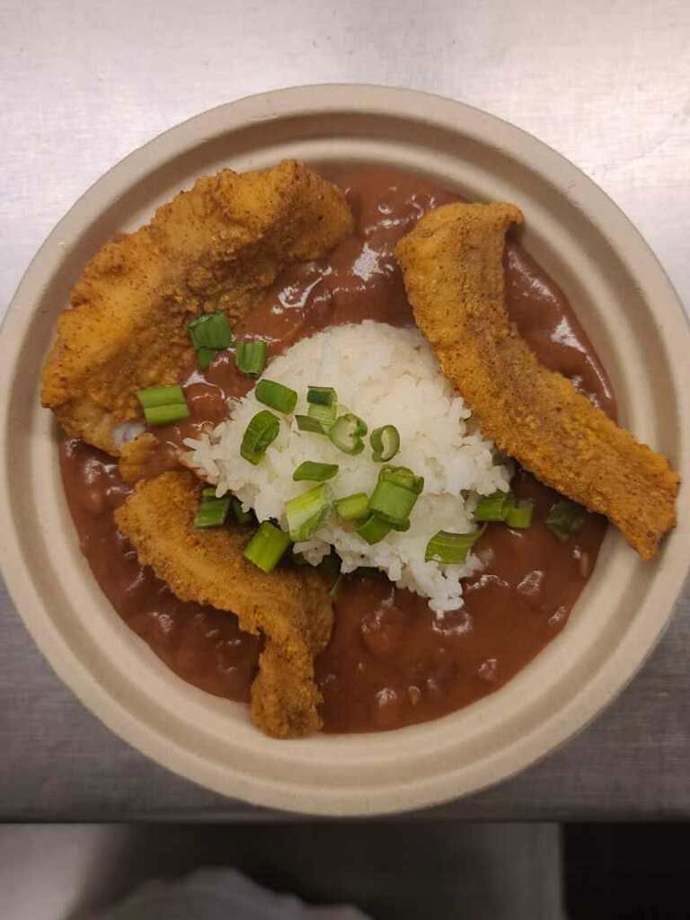 A fried catfish dish over gumbo in a brown compostable takeout bowl