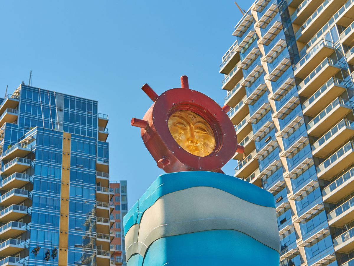 A sculpture of a face upon a ship’s wheel in front of two modern residential towers.