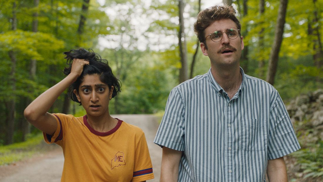 Actors Sunita Mani and John Reynolds stand on a narrow road through the woods looking frazzled in the alien invasion comedy Save Yourselves!