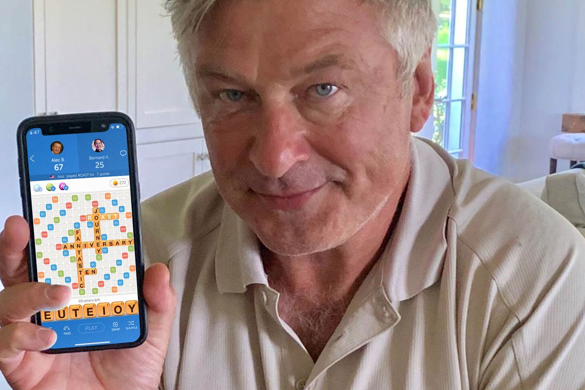 An aging man — Alec Baldwin&nbsp;— with gray hair and a tan polo shirt has a mostly neutral expression on his face. He is slightly smirking and holding up an iPhone with a game of Words With Friends on the screen.