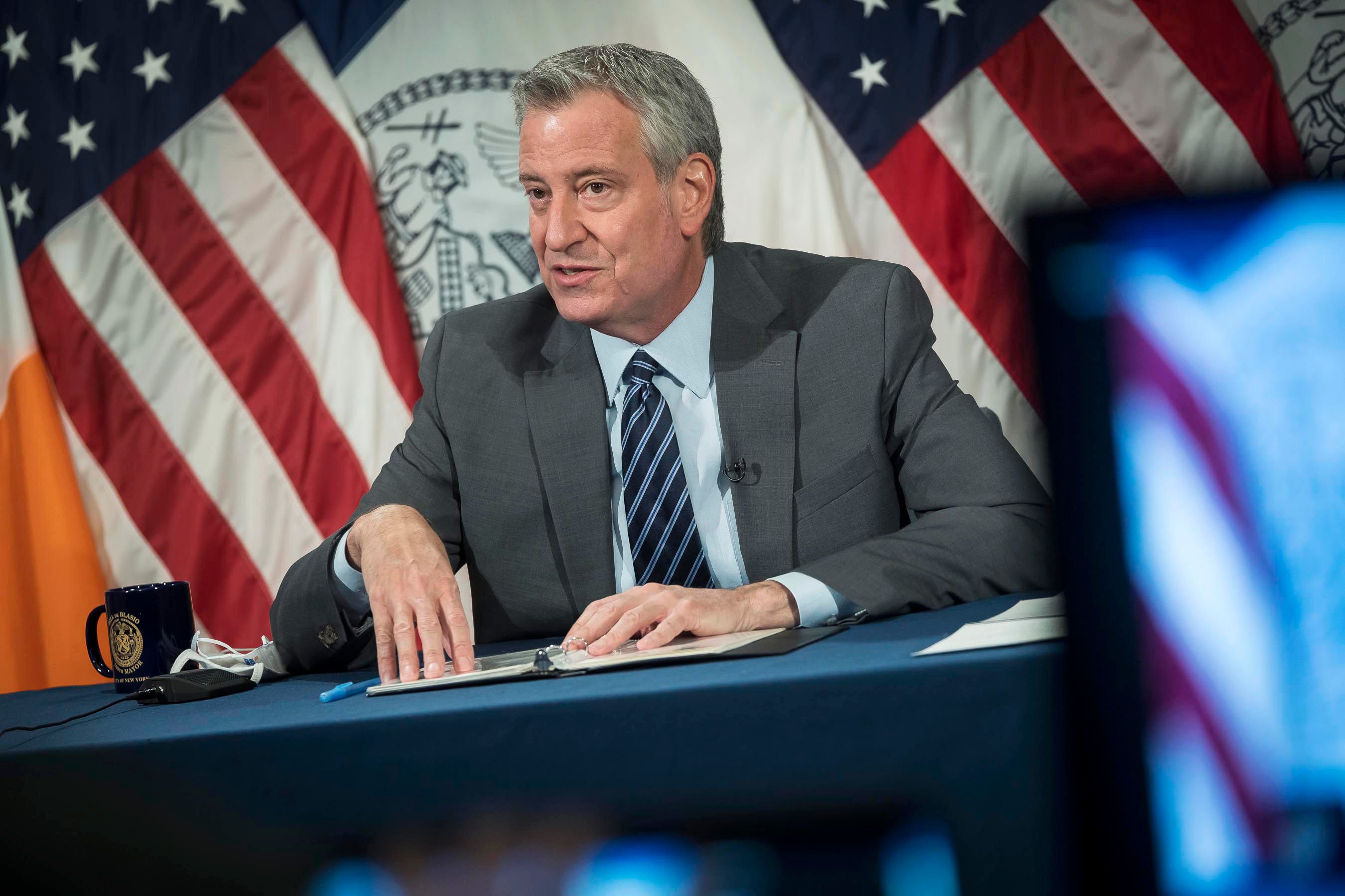 Mayor Bill de Blasio talks while sitting at a desk with U.S. and NYC flags behind him.