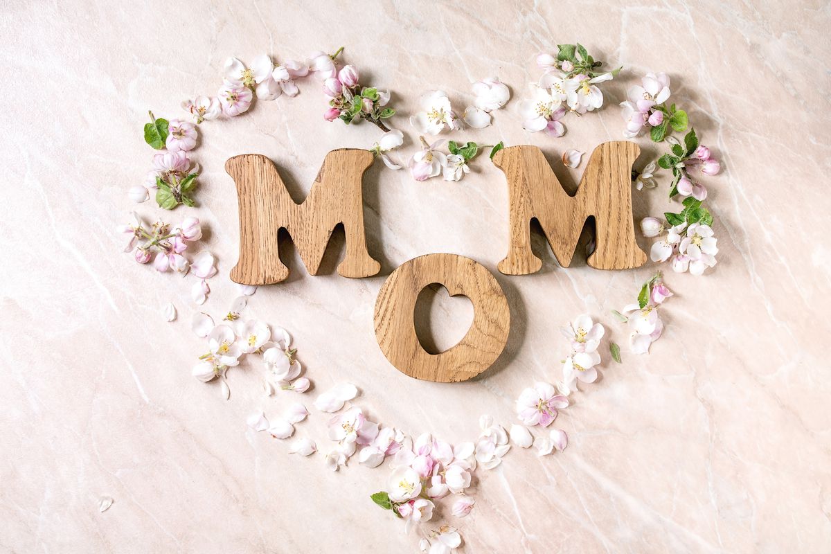 Mother’s day greeting card. Wooden letters MOM with spring apple flowers and petals as heart shape over pink marble background. Flat lay, space