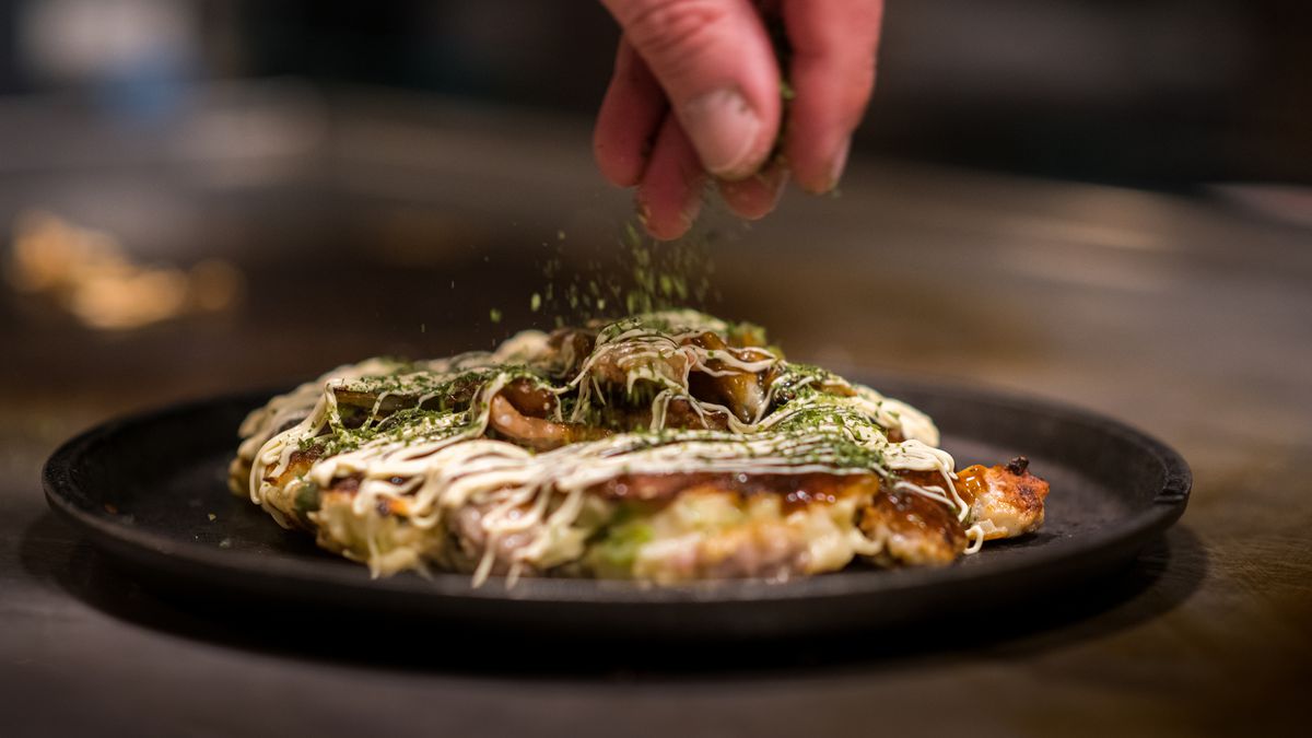 A black round plate with okonomiyaki, with a hand out of frame sprinkling seasoning.