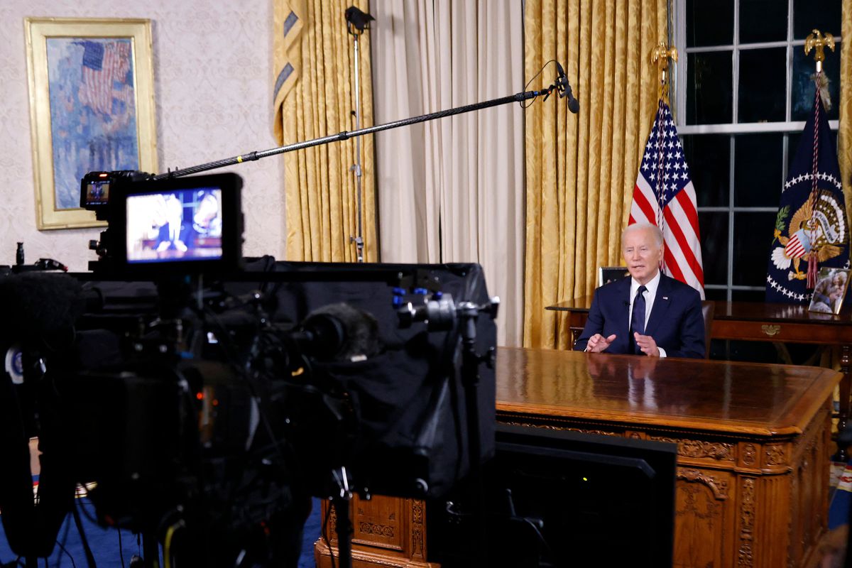 Biden sits at a desk with a video monitor visible in the front of the frame. 