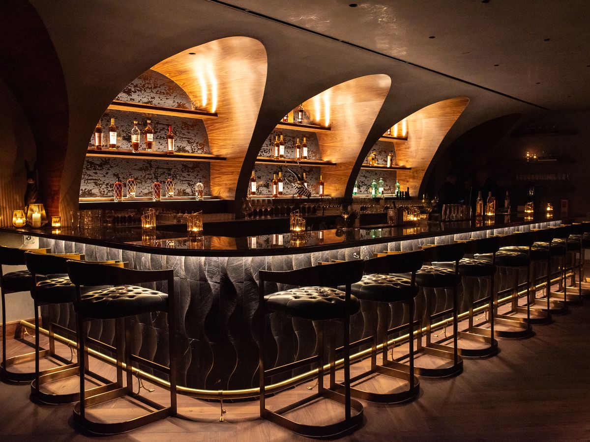 An illuminated bar, outfitted with sleek shelves and surrounded by bar seats,