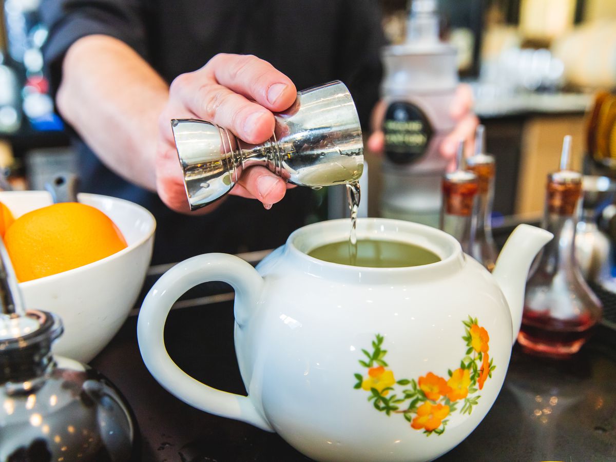 A bartender pours alcohol out of a jigger into a white ceramic teapot with orange flowers on the side.