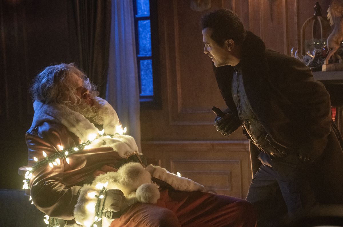 Scrooge (John Leguizamo) leans menacingly over Santa Claus (David Harbour), who’s tied to a chair with a string of lit-up white Christmas lights, in Violent Night