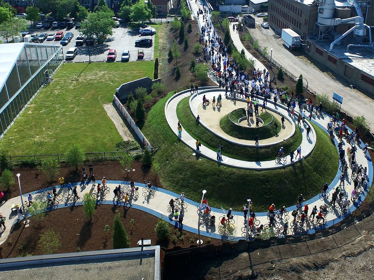 A circular bike trail on a hill. There are multitudes of people riding bicycles along the trail. The trail is surrounded by a large green grass lawn. 