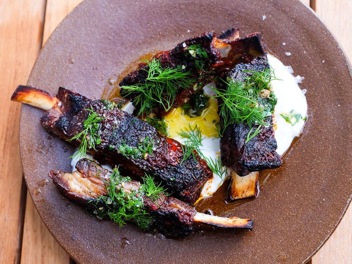 Four ribs on a brown stoneware plate on a bed of creamy yogurt with sprigs of bright green herbs on top. 