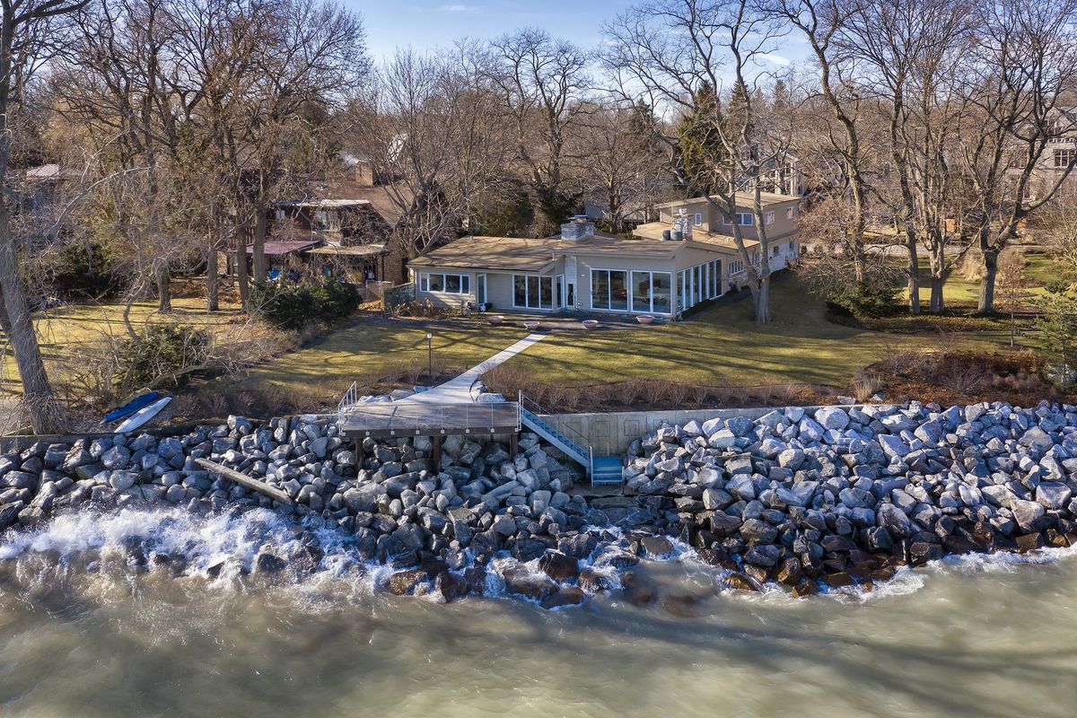 An aerial image of a ranch home on a lake with large stones forming a breakwater. There are stairs down to the shoreline. 