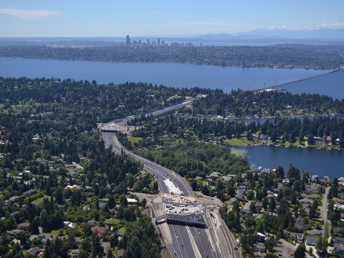 A highway winds through a wooded area, eventually turning into a bridge over a large lake. Skyscrapers are visible in the background.