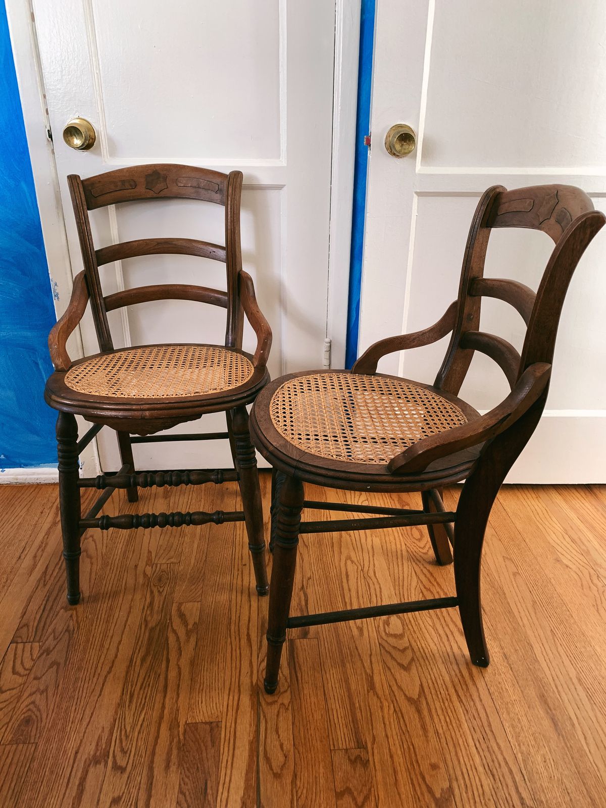Two wooden chairs with woven bottoms are placed next to each other in a room that has light blue walls, two white doors, and wooden floors.