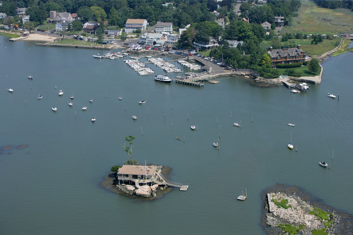 An aerial view of a house on an island with boats scattered in the water. A small town and marina are just off the island. 