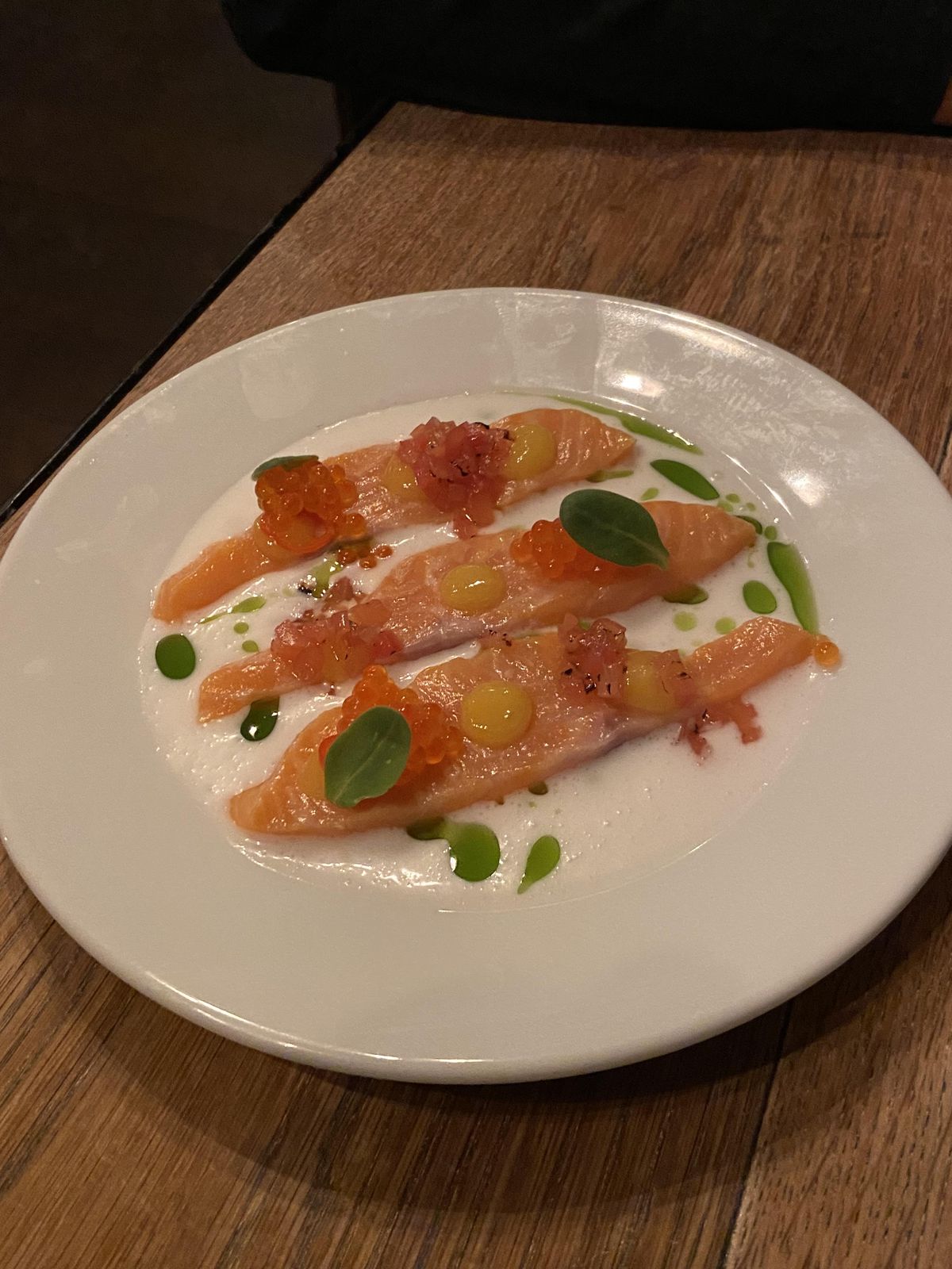 Three thick slices of cured trout on a white plate, with coconut sauce, a herb oil, and trout roe
