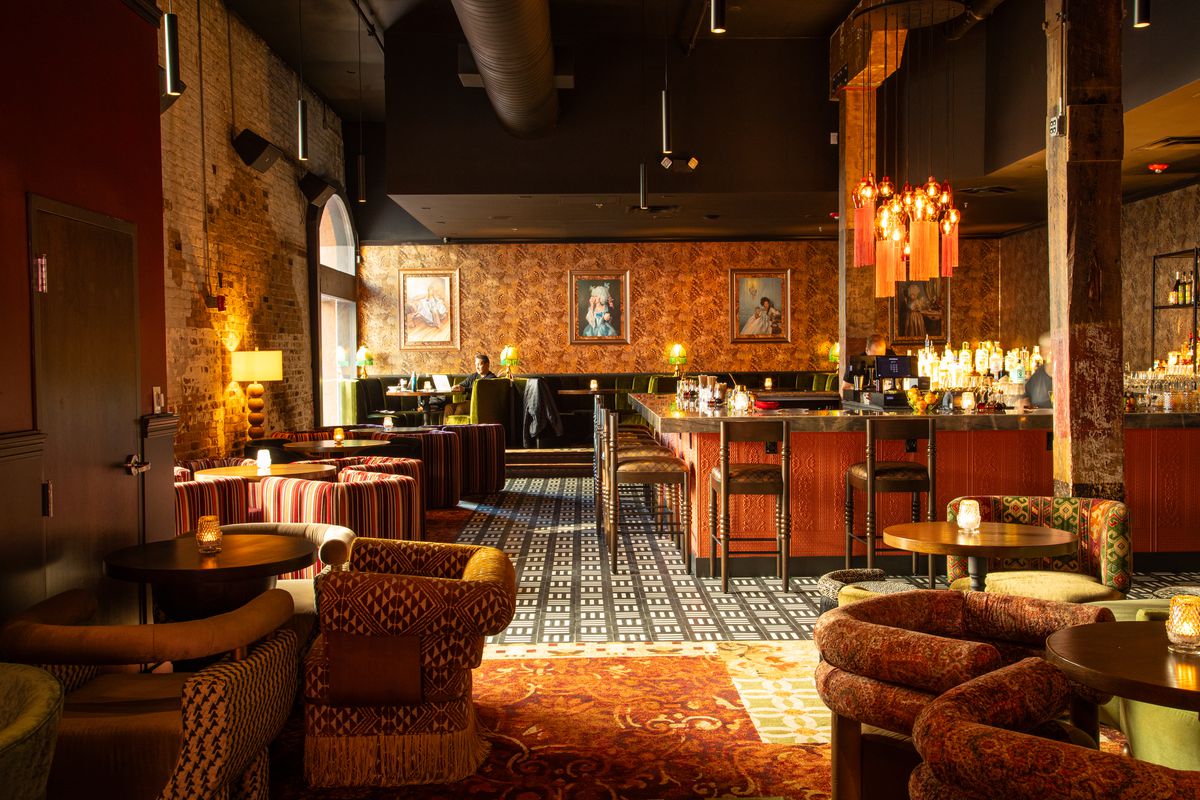 A large bar and lounge space with low-slung arm chairs, patterned wallpaper and flooring, and an L-shaped bar. 