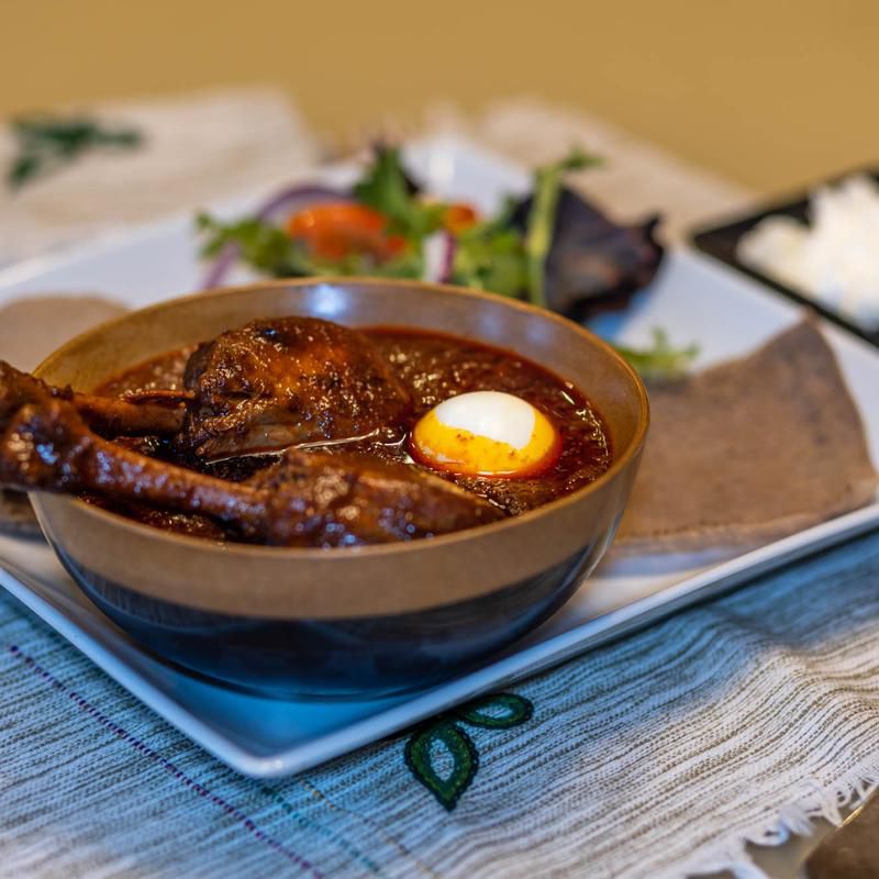 A bowl of rich meat stew with an egg on it on a white plate, with injera bread and salad in the background. 