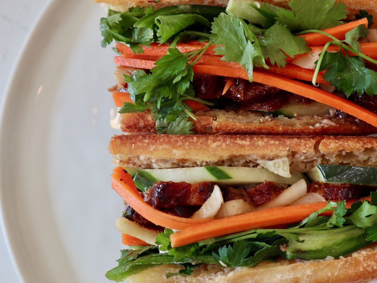 overhead shot of banh mi sandwich, close up on right side of a white plate .topped with shredded carrots, cilantro, and onions.