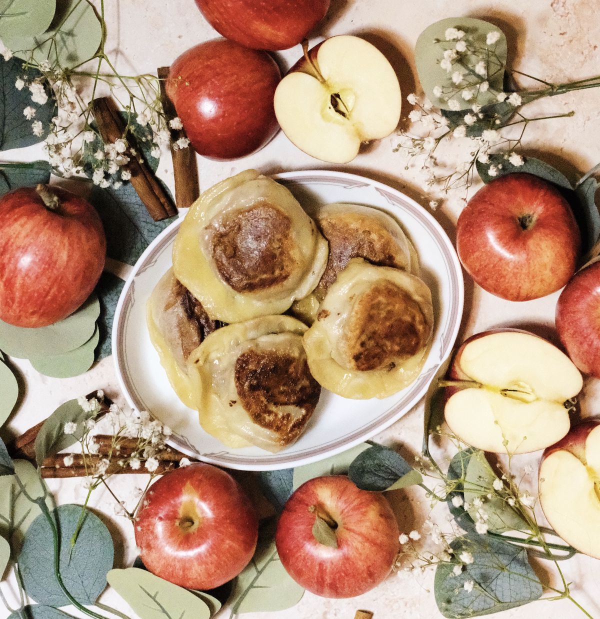 Five pierogi on a white oval plate, several whole and sliced red apples, and some foliage
