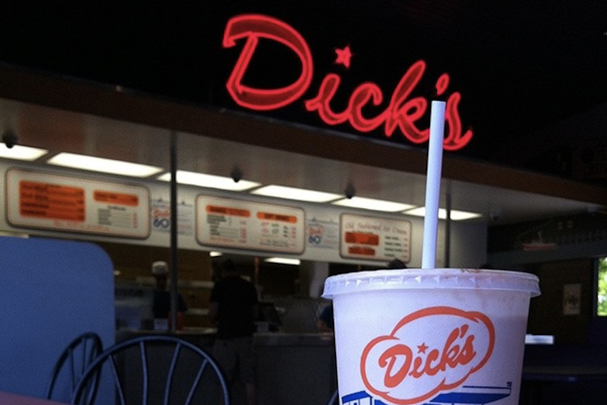 The storefront of Dick’s Drive-In with its red neon sign lit up and a Dick’s-labeled paper cup in the foreground