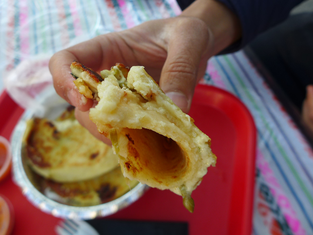 A hand holds a folded pupusa, whose glistening inside looks corn yellow and still chewy. In the background, a red takeout tray is visible.