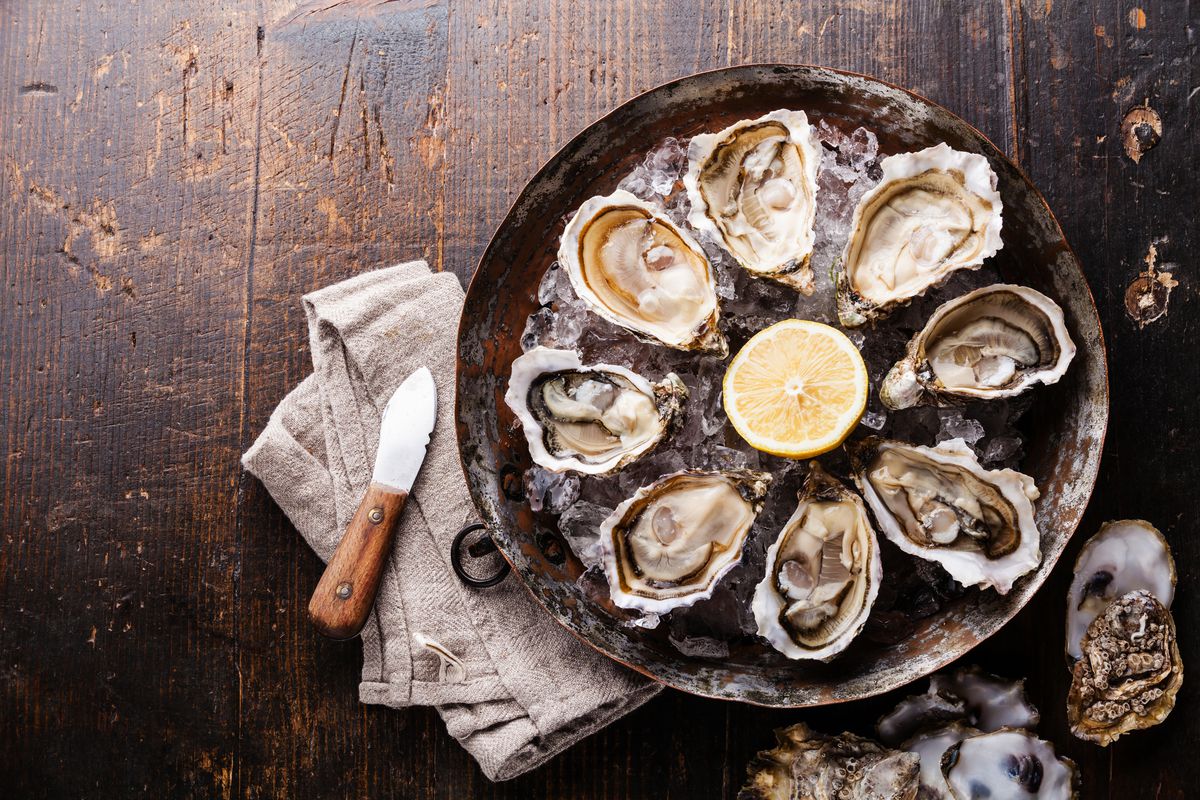 Stock photograph of oysters. An overhead view shows a platter of eight oysters on the half shell on ice, with a sliced lemon in the middle, all sitting on a dark wooden table.