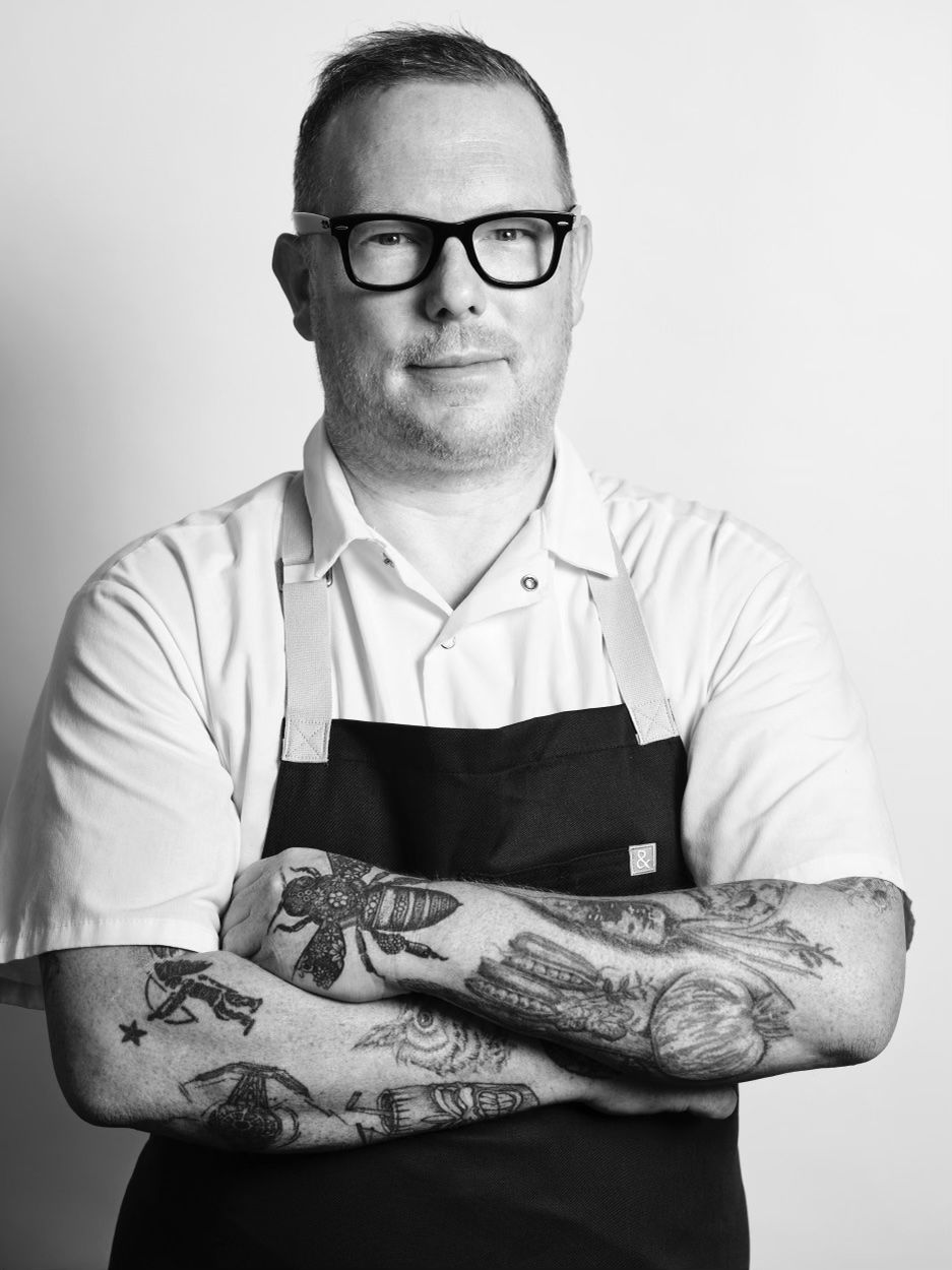 Tattooed chef wearing chef coat and black apron stands folding his arms.
