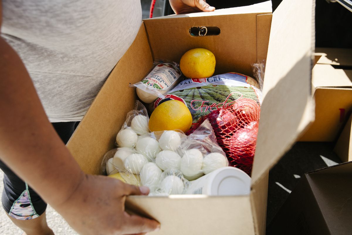 A person carries a cardboard box containing eggs, potatoes, cheese, and other food items.