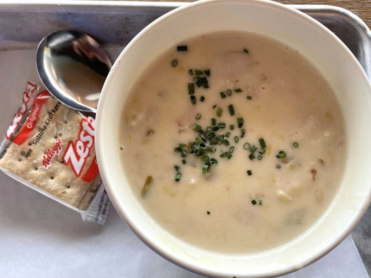 A bowl of creamy, white clam chowder topped with diced chives, sits next to a little pack of saltines.
