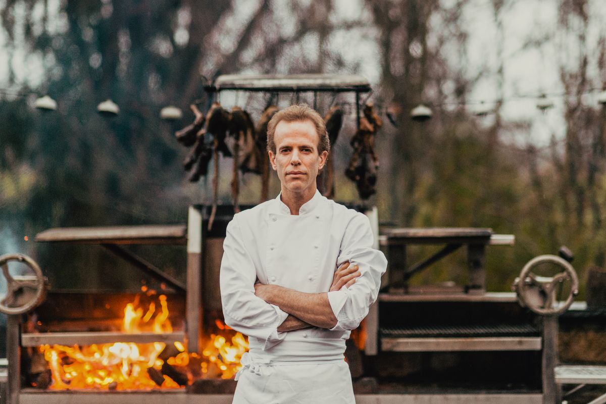 Dan Barber stands outside at Blue Hill at Stone Barns with his arms crossed and a fire roaring behind him