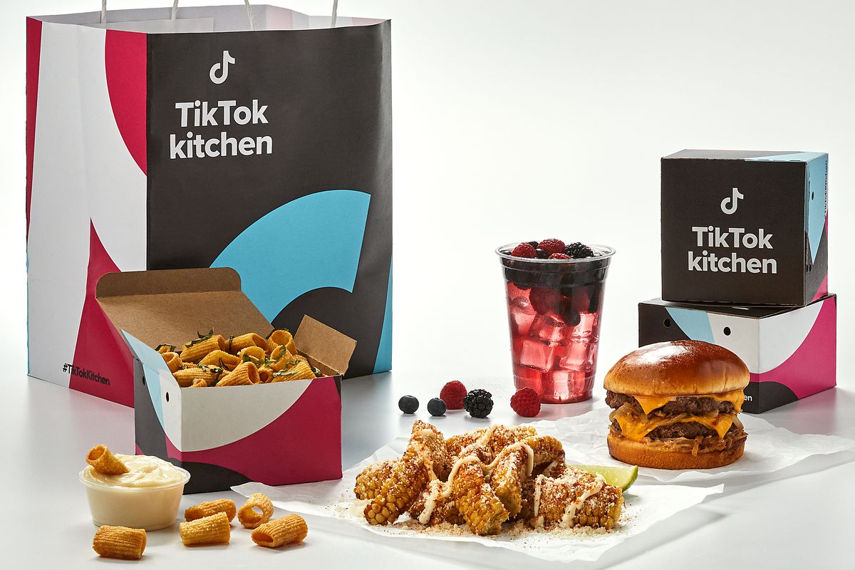 Delivery bags with the name “TikTok Kitchen” behind delivery food, including a double cheeseburger, corn ribs, and pasta chips.