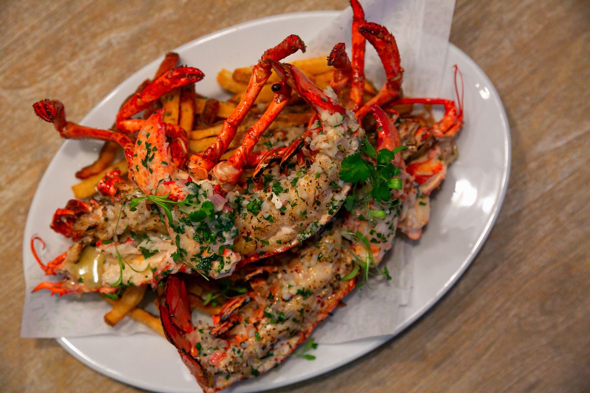 A cooked lobster garnished with green herbs sits on an oval white plate with a side of fries.
