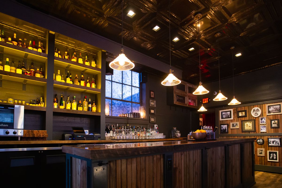 A dark wooden bar with six vintage lights hanging above it and a back bar lit with bottles of liquor on the shelves. The light is reflecting off the tin ceiling above the bar