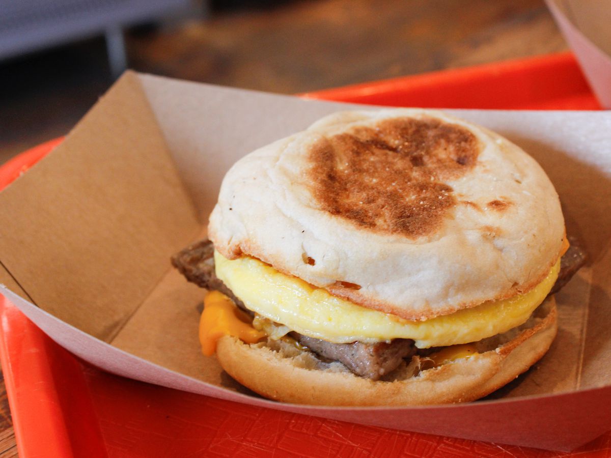 A sausage, egg, and cheese breakfast sandwich sits in a paper container on a red tray on a wooden table