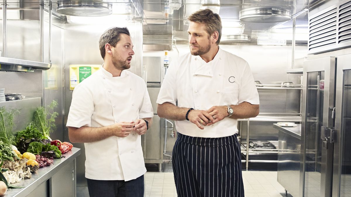 Curtis Stone and Christian Dortch talk while walking through a kitchen in chef’s clothes.