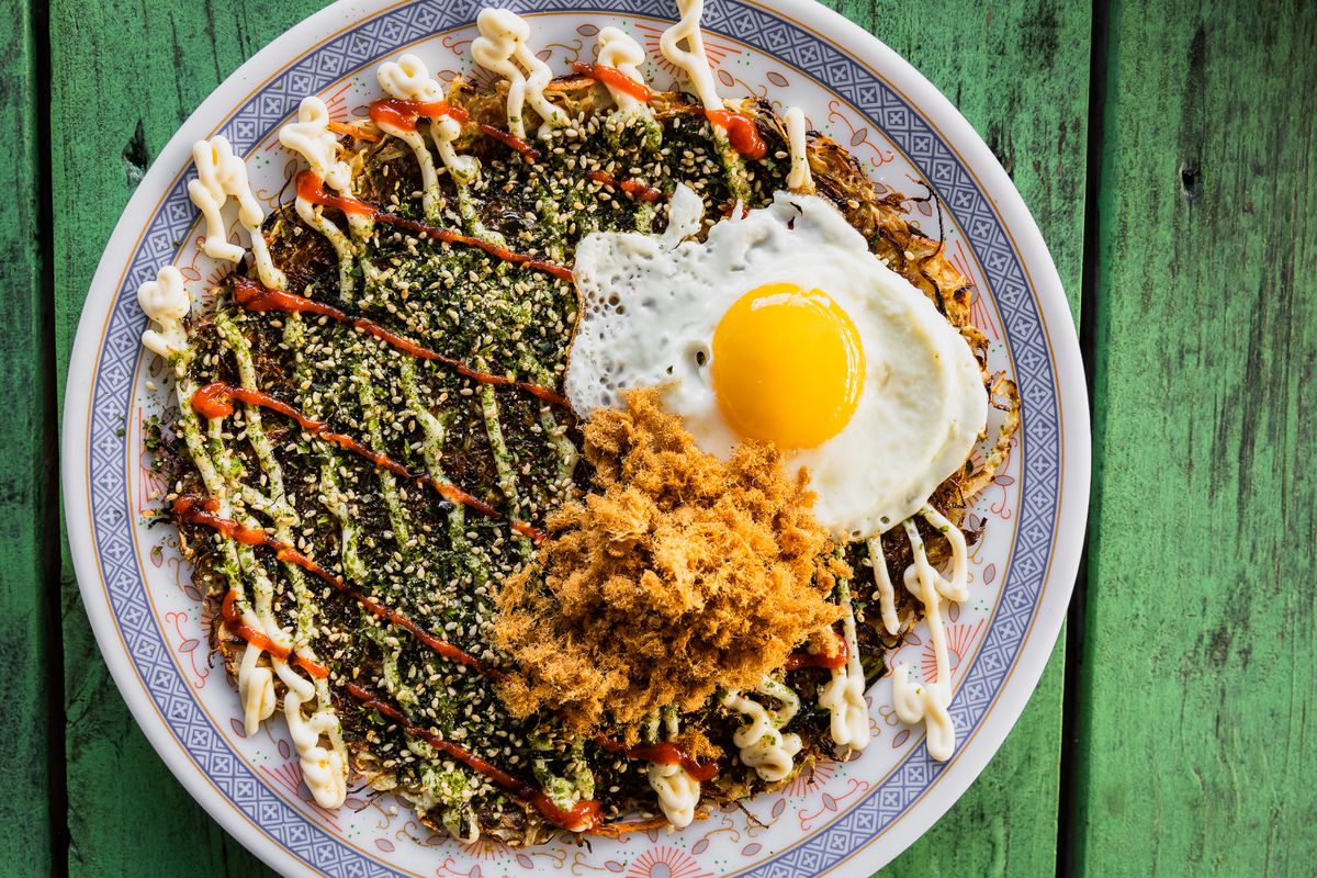 A top-down view of a Japanese okonomiyaki topped with pork candy, a sunny side up egg, stripes of mayo and a red sauce, and lots of seasoning