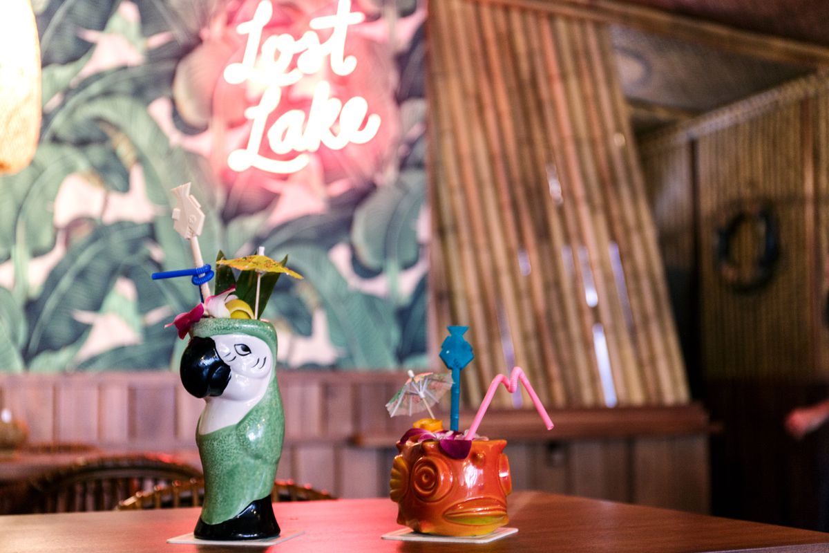 Two drinks with umbrellas and colorful straws on a bar, one in a glass shaped like a parrot, the other a fish. Tropical wallpaper and neon sign in the background.