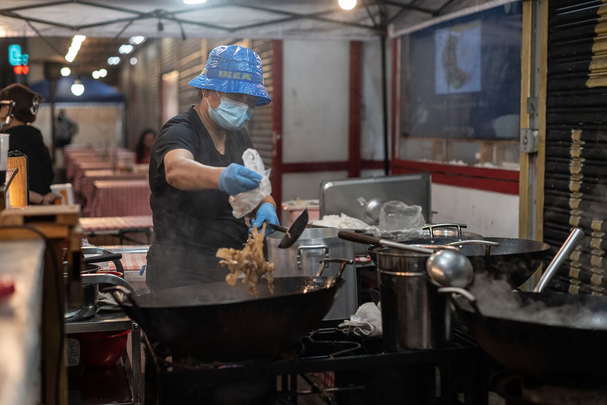 A woman wearing a hat made of blue IKEA bag material prepares a Thai dish over a wok at Rad Nah Silom in East Hollywood, California.