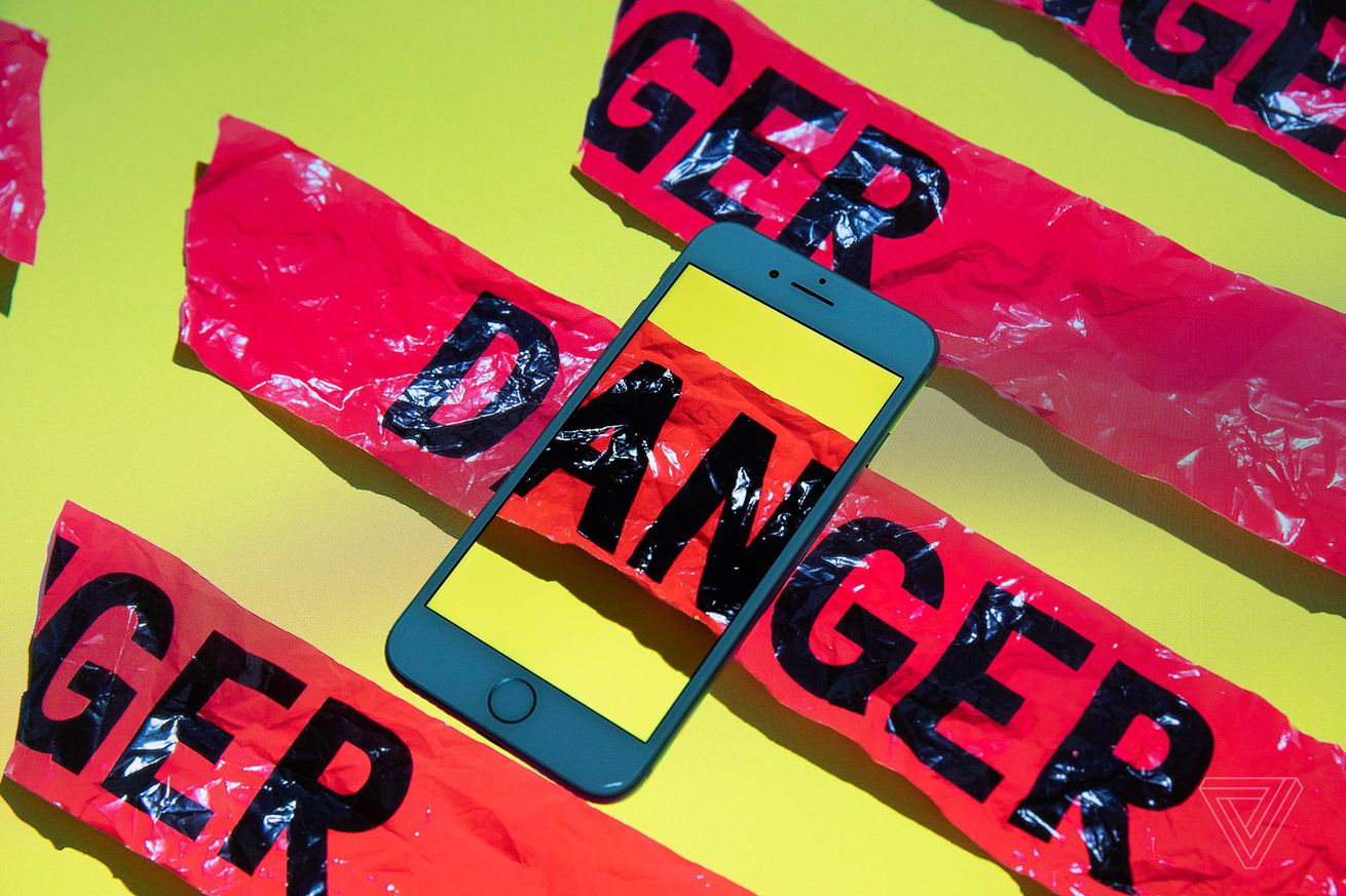 A smartphone sits on top of a surface with red tape reading “DANGER.” Where one strip intersects the phone, it continues inside the phone’s screen.