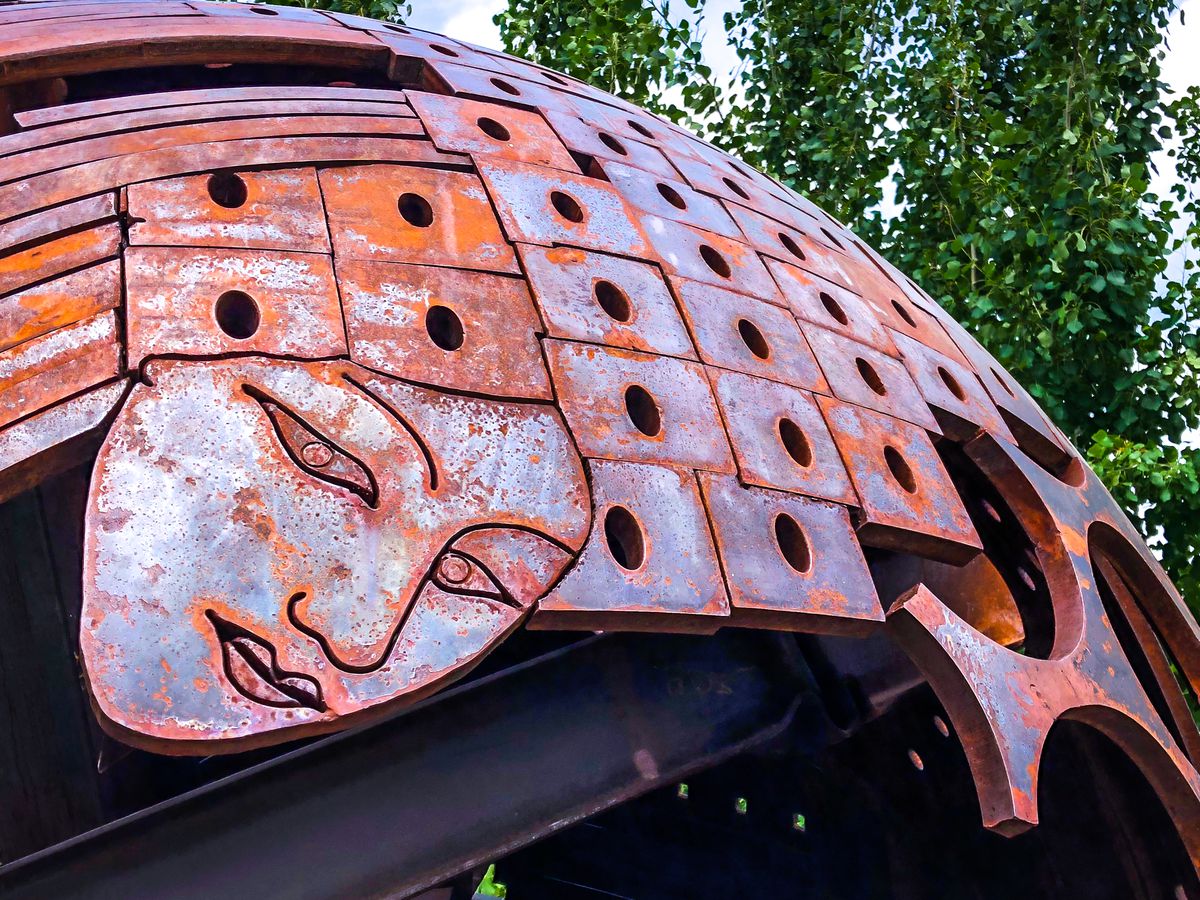 A dome-shaped sculpture made of rusty, metal, rectangle tiles with holes through the center of each. At the bottom left, a metal shape has a feminine face etched in simple line art.