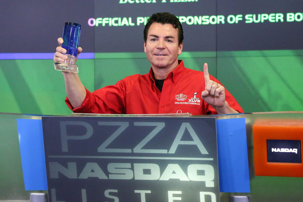 John Schnatter, formerly of Papa John’s, wears a red zip-up and stands in front of blue and green graphics when ringing the opening bell at NASDAQ.