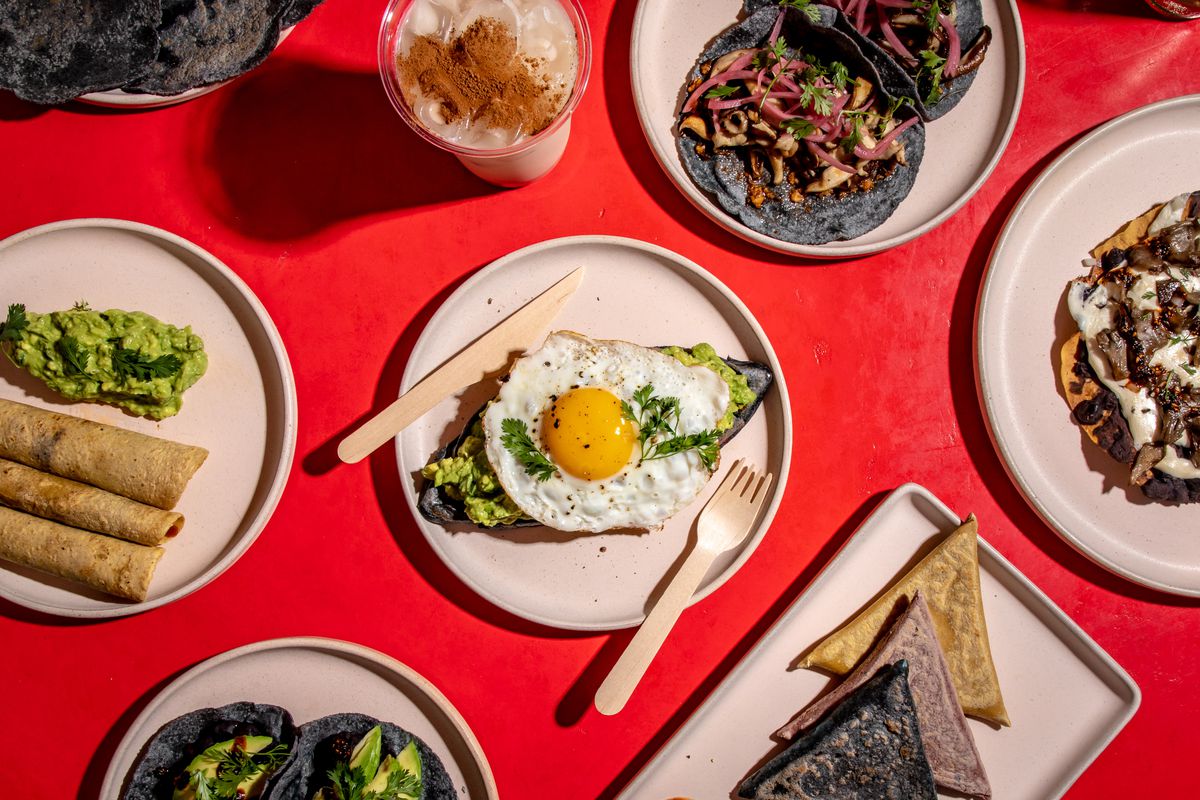 A sunny-side up fried egg sits over a memela with avocado in an overhead shot; the memela is surrounded by assorted dishes including tacos on blue corn tortillas and a cup of horchata